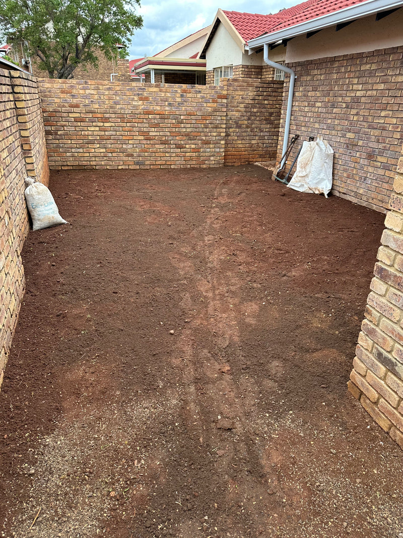 A freshly landscaped backyard or patio area with reddish-brown soil cleared and leveled, bordered by a brick wall on the left, a brick wall on the right, and a brick fence in the background. Two white sandbags sit on the left side, and a shovel and white sack are leaning against the right wall.